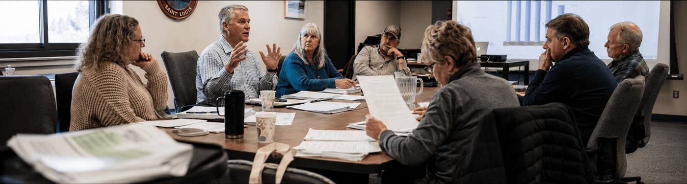 A county workforce board mid-discussion around a conference table, papers spread, a chart projected behind them